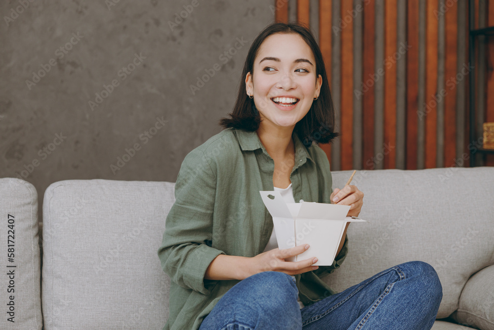 Young woman of Asian ethnicity wearing casual clothes eat Chinese food cuisine hold takeaway container box sits on grey sofa couch stay home hotel flat rest relax spend free spare time in living room. Young woman of Asian ethnicity wearing casual clothes eat Chinese food cuisine hold takeaway container box sits on grey sofa couch stay home hotel flat rest relax spend free spare time in living room.