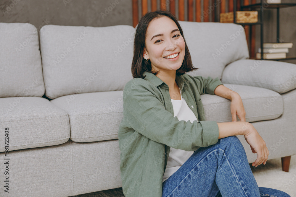 Young smiling happy cool fun satisfied woman of Asian ethnicity wears casual clothes look aside sit near grey sofa couch stay at home hotel flat rest relax spend free spare time in living room indoor Young smiling happy cool fun satisfied woman of Asian ethnicity wears casual clothes look aside sit near grey sofa couch stay at home hotel flat rest relax spend free spare time in living room indoor
