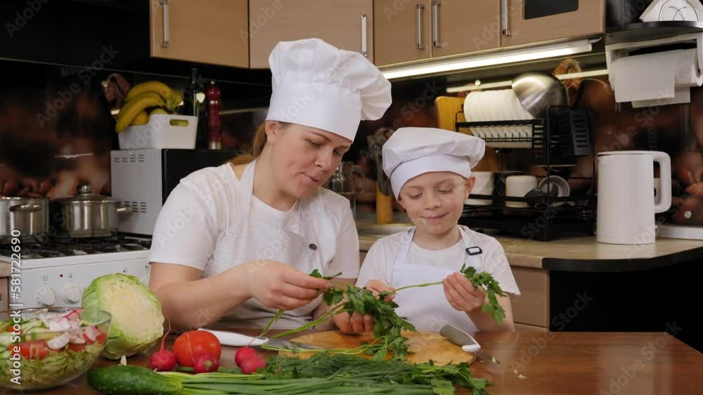 A mother with her little son in aprons and caps are preparing a salad together in the kitchen, they are sniffing parsley and chopping it. A mother with her little son in aprons and caps are preparing a salad together in the kitchen, they are sniffing parsley and chopping it.