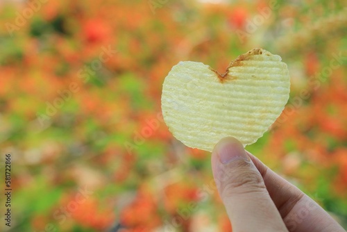 Heart-shaped potato chip in woman hand with colorful leaves in autumn background. Food and Valentine's day concept.
