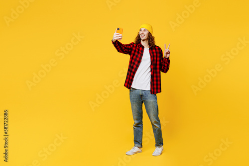 Full body young man wearing red checkered shirt white t-shirt hat doing selfie shot on mobile cell phone post photo on social network show v-sign isolated on plain yellow background studio portrait. © ViDi Studio