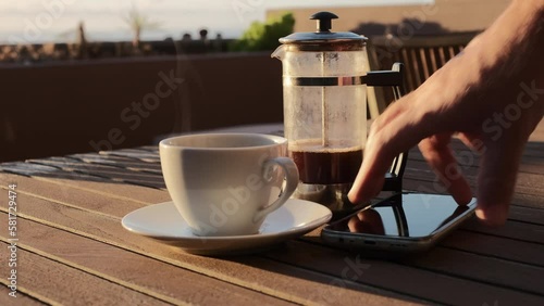 Café caliente humeante de cafetera de émbolo sobre una mesa de madera con luz de amanecer en una terraza con vistas al mar y jardinera de fondo, con mano de hombre que coge un móvil y unas gafas.