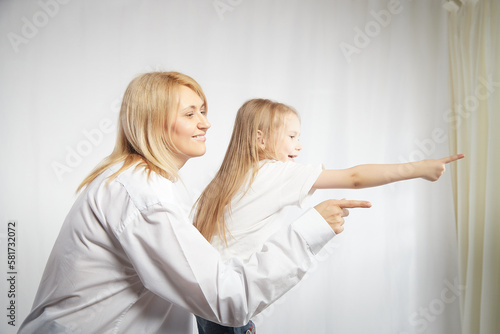 Portrait of a blonde mother and daughter who having communicate and play on a white background. Mom and little girl models pointing his finger forward. The concept of family unity