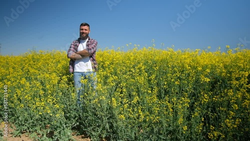 Happy and successful farmer is standing by his rapeseed field. Rapeseed plantation in bloom. Slow motion video.