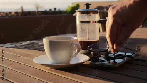 Café caliente humeante de cafetera de émbolo sobre una mesa de madera con luz de amanecer en una terraza con vistas al mar y jardinera de fondo, con mano de hombre que coloca un móvil y unas gafas.