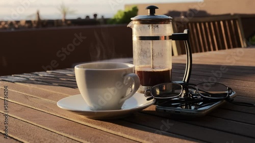 Café caliente humeante de cafetera de émbolo sobre una mesa de madera con luz de amanecer en una terraza con vistas al mar y jardinera de fondo.