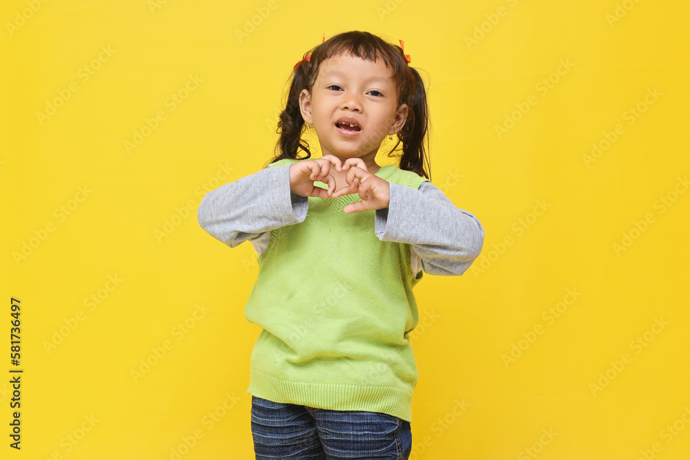 Attractive Asian little girl smiling to the camera while finger make a heart shape isolated on yellow background. Attractive Asian little girl smiling to the camera while finger make a heart shape isolated on yellow background.