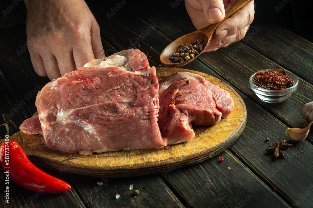 The hands of the cook add pepper to raw meat before grilling. Preparing beef meat before roasting. Working environment in kitchen of a restaurant or hotel