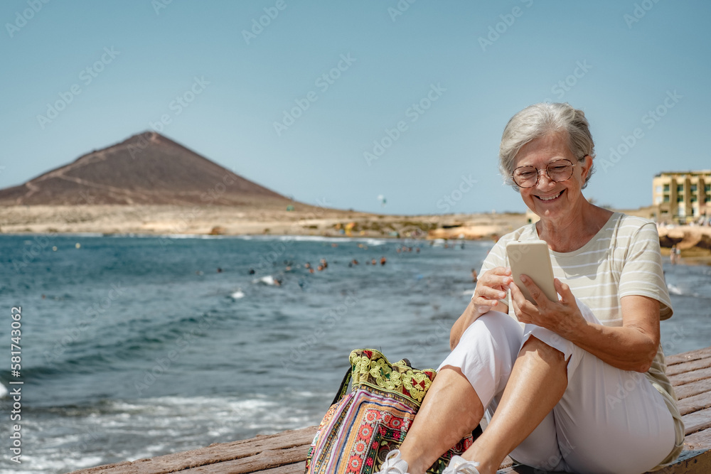 Video call concept. Smiling senior woman using smartphone for online communication via video meeting sitting at the beach, happy grey-haired woman looking and talking into phone webcam