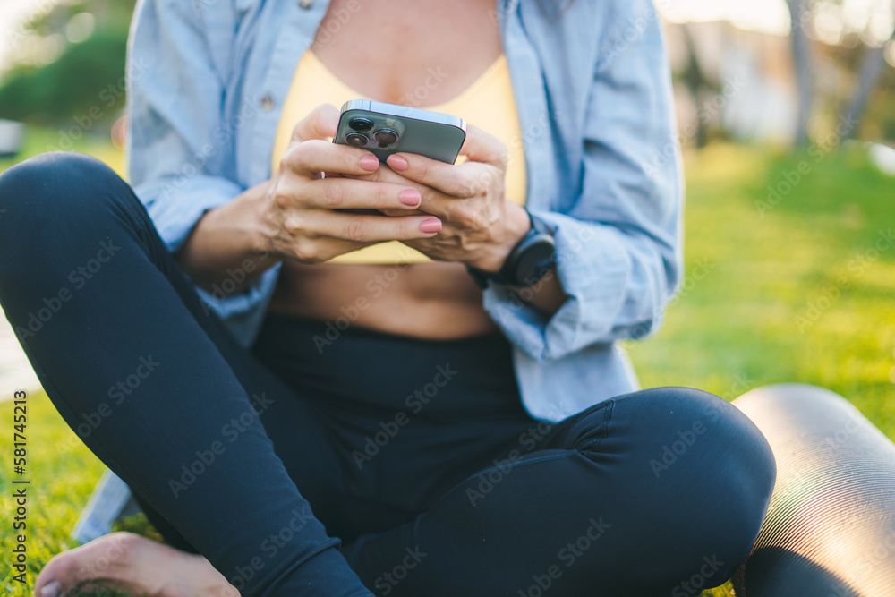 Fitness woman using phone application for sport and workout, sitting on grass with rubber yoga mat in park after training session. Happy lifestyle. Sitting