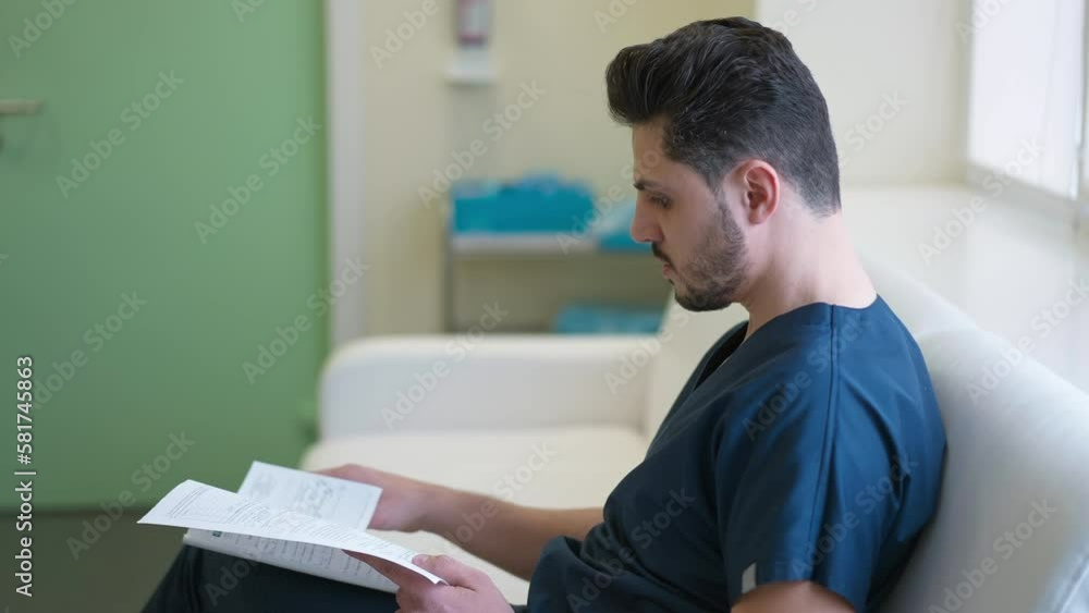 Side view portrait of focused intelligent surgeon examining case history sitting in hospital hallway. Smart concentrated Middle Eastern young handsome man in uniform indoors in medical clinic Side view portrait of focused intelligent surgeon examining case history sitting in hospital hallway. Smart concentrated Middle Eastern young handsome man in uniform indoors in medical clinic