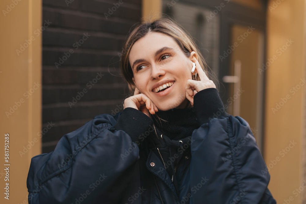 Pensive lady in stylish bomber enjoying good autumn or spring weather. Outdoor portrait of blonde girl in white wireless earphones listening music during walk on the street in morning. Pensive lady in stylish bomber enjoying good autumn or spring weather. Outdoor portrait of blonde girl in white wireless earphones listening music during walk on the street in morning.