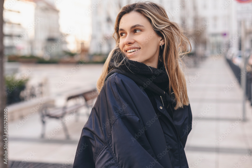 Happy blonde woman wear navy blue bomber jacket walking in the street and smiling to camera. Stylish girl in fashion outfit walking over the city. What a beautiful day out. Girl turn around.