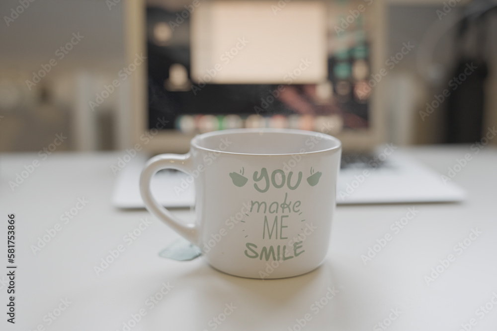 Focus on a labeled cup of coffee in the foreground. In the blurred background is an open laptop with open tabs on a desk Focus on a labeled cup of coffee in the foreground. In the blurred background is an open laptop with open tabs on a desk