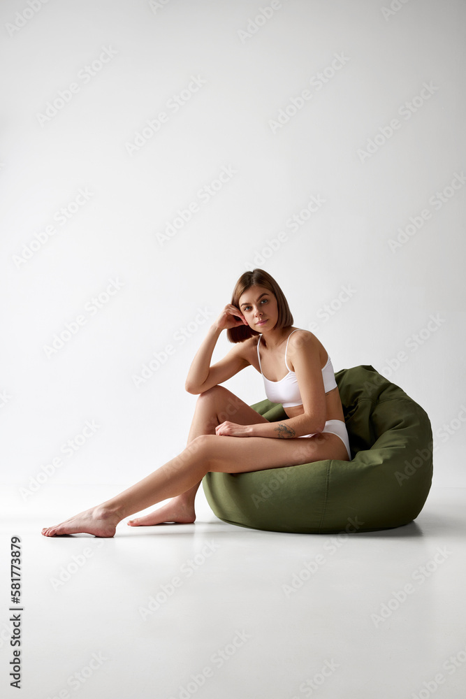 Beautiful woman sitting on big pillow chair in white underwear isolated over grey background. Sportive body. Concept of beauty, skin care, health, cosmetics, relax time Beautiful woman sitting on big pillow chair in white underwear isolated over grey background. Sportive body. Concept of beauty, skin care, health, cosmetics, relax time
