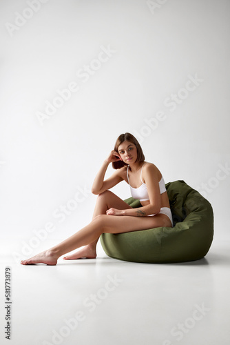 Beautiful woman sitting on big pillow chair in white underwear isolated over grey background. Sportive body. Concept of beauty, skin care, health, cosmetics, relax time