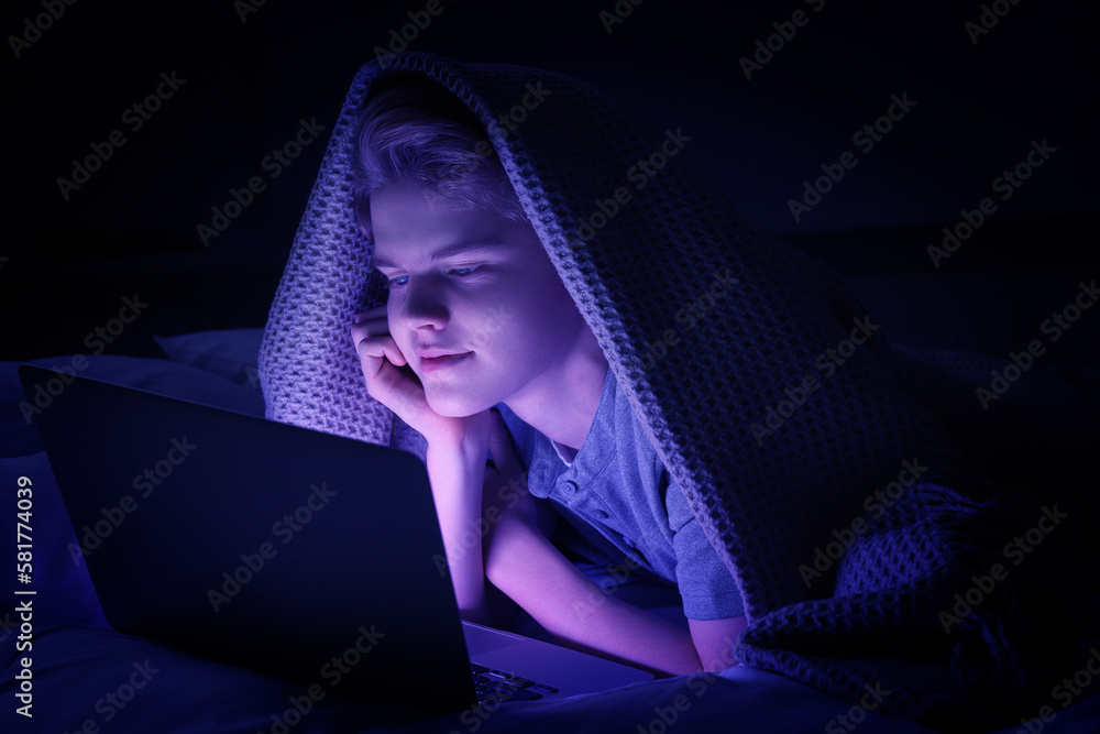 Internet addiction. Teenage boy using laptop under blanket on bed at night. Toned in blue Internet addiction. Teenage boy using laptop under blanket on bed at night. Toned in blue