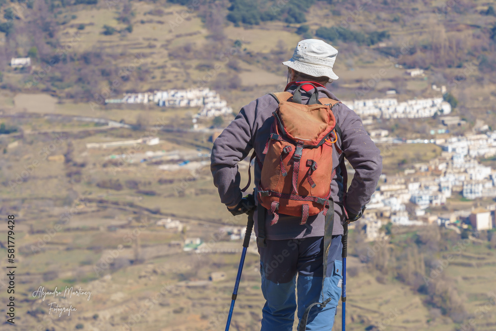 A man at the top of a mountain wearing a hat and carrying hiking poles.