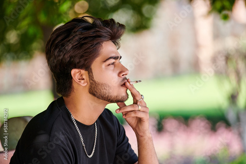 Bild auf Leinwand Young indian man smoker portrait in black t shirt and silver neck chain in public park, hindu male smoking close up portrait