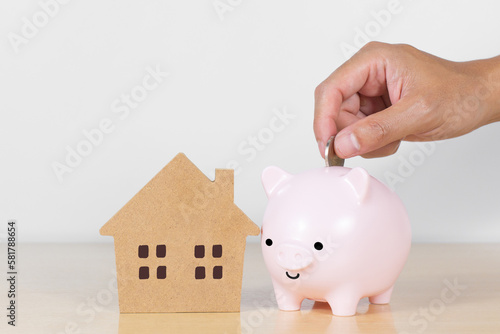 Hand inserting coin inside of piggy bank with house model on wooden table. Concept for invest, residential investment and saving success in the future goal