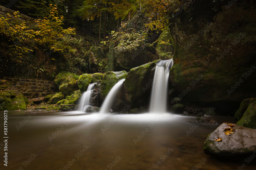 Obraz premium Horizontal view on a wet rock in a lake with Schiessentümpel waterfalls and trees in the background. Autumn landscape in Müllerthal with long exposure and copy space. Petite Suisse