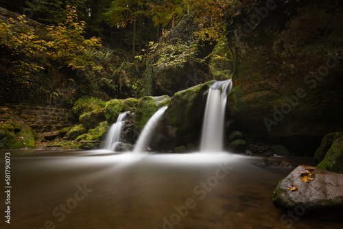 Horizontal view on a wet rock in a lake with Schiessentümpel waterfalls and trees in the background. Autumn landscape in Müllerthal with long exposure and copy space. Petite Suisse