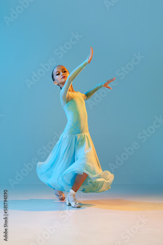 Valse. Portrait of little flexible girl in stage outfit, dress dancing ballroom dance over blue background. Concept of beauty, professional dances, skills