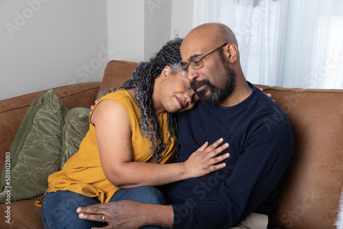 Portrait of a middle aged black married couple sitting on the couch. The wife is comforting her husband while he is distressed. 