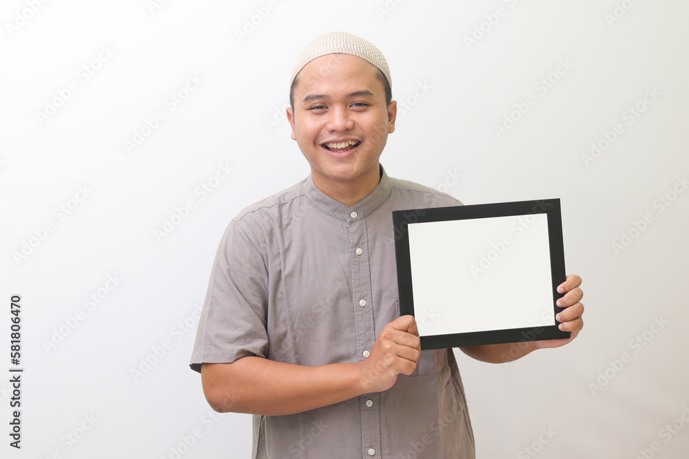 The portrait of the religious wearing a muslim gray shirt and a white hat smiled broadly holding a whiteboard. Isolated image on white background The portrait of the religious wearing a muslim gray shirt and a white hat smiled broadly holding a whiteboard. Isolated image on white background