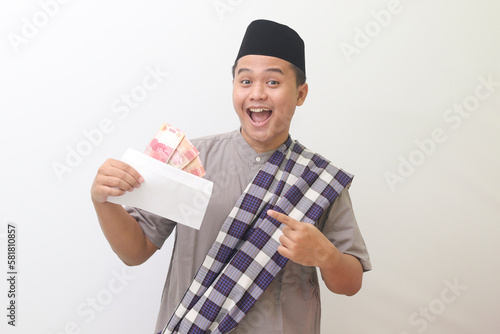 Portrait of smiling Asian man smiling happily at camera wearing gray muslim shirt and black hat with sarong slung over shoulder pointing to envelope filled with money he is carrying. Isolated image on photo