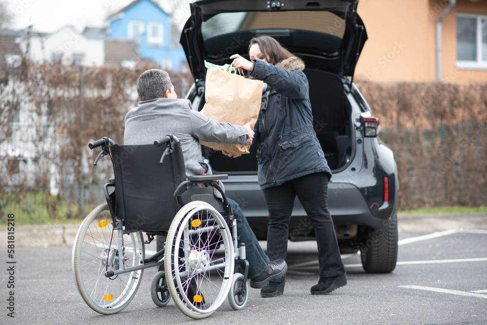 A woman helps a disabled person on a wheelchair to pack groceries into the car