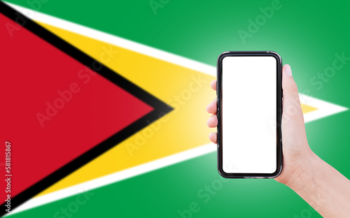 Male hand holding smartphone with blank on screen, on background of blurred flag of Guyana. Close-up view.