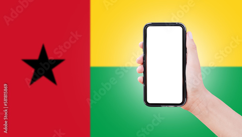 Male hand holding smartphone with blank on screen, on background of blurred flag of Guinea-Bissau. Close-up view.