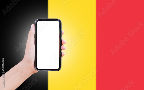 Male hand holding smartphone with blank on screen, on background of blurred flag of Belgium. Close-up view.
