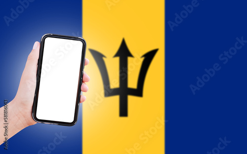 Male hand holding smartphone with blank on screen, on background of blurred flag of Barbados. Close-up view.