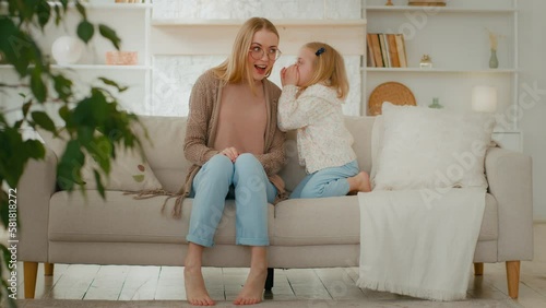 Caucasian little girl daughter child kid whisper secret gossip to mum share secrecy information honestly talking to mommy surprised amazed shocked woman mother babysitter smile listen sitting at couch photo