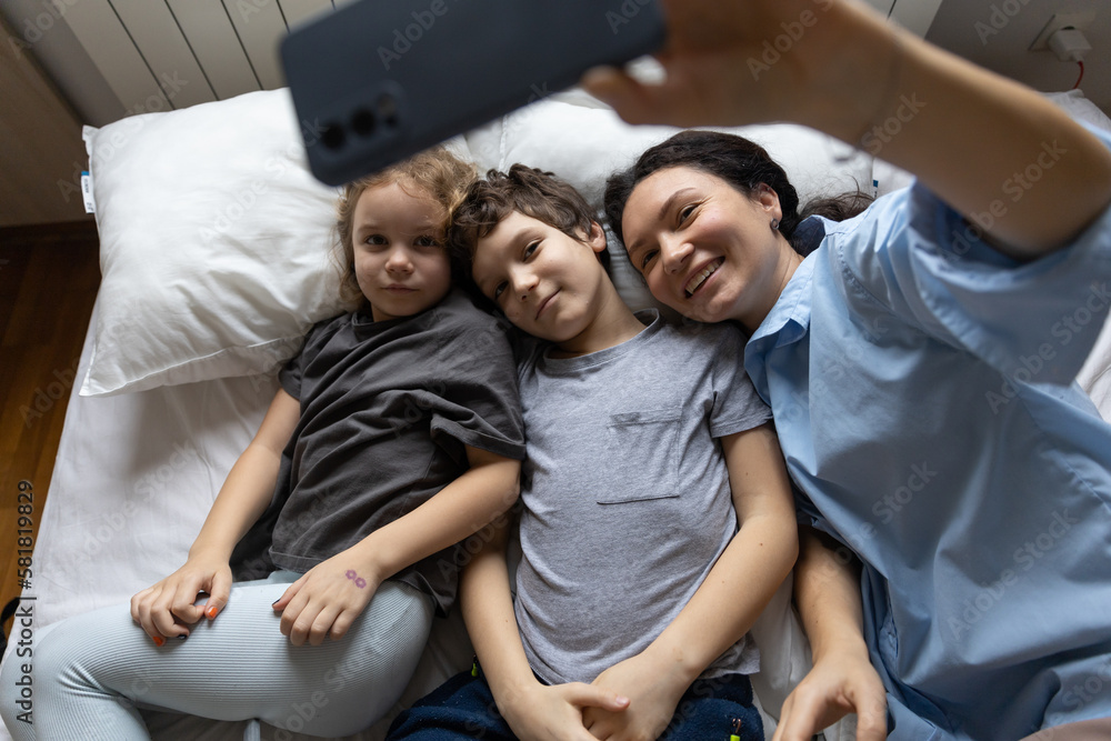 Fototapeta premium Happy caucasian mother and 2 kids have online video call with father or grandmother mobile phone when lying on bed at home. the concept of modern communication.lifestile. top view