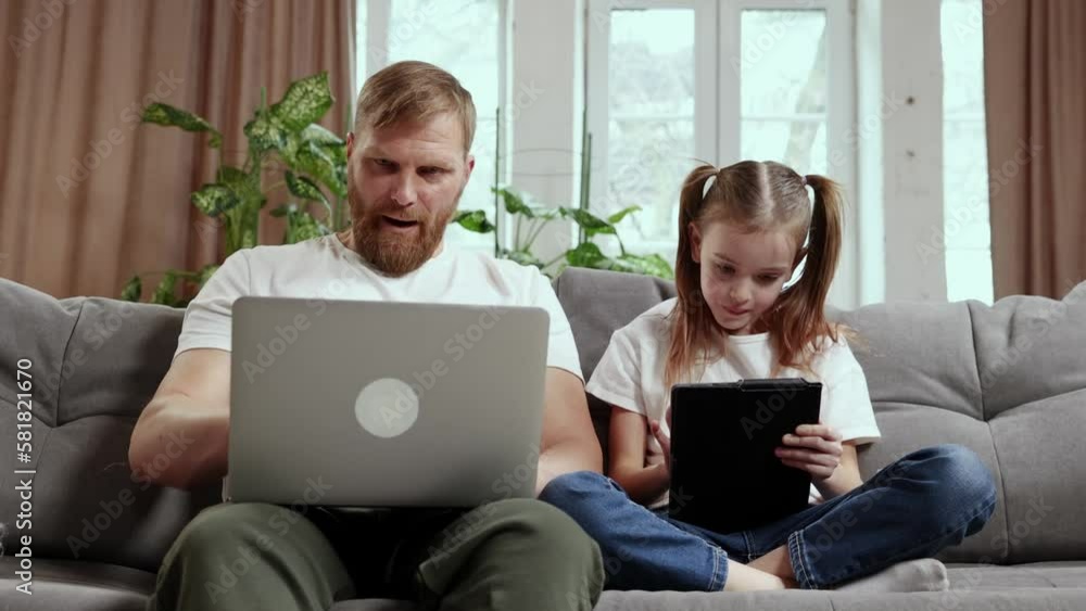 Family time. Mature bearded man, father working on laptop remotely at home, sitting on couch with his little girl daughter playing on tablet. Fatherhood, childhood, freelance job, remote work concept Family time. Mature bearded man, father working on laptop remotely at home, sitting on couch with his little girl daughter playing on tablet. Fatherhood, childhood, freelance job, remote work concept