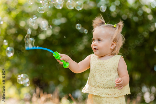 a happy little blonde girl in a yellow dress blows soap bubbles in the park in summer. Earth Day. Children Protection Day