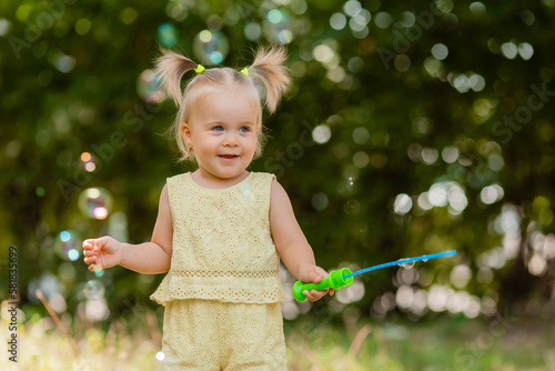 a happy little blonde girl in a yellow dress blows soap bubbles in the park in summer. Earth Day. Children Protection Day