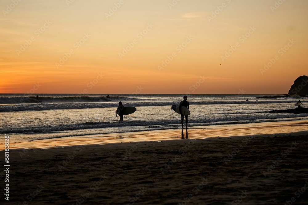 People walking along a sandy beach, each carrying a long surfboard in their hands