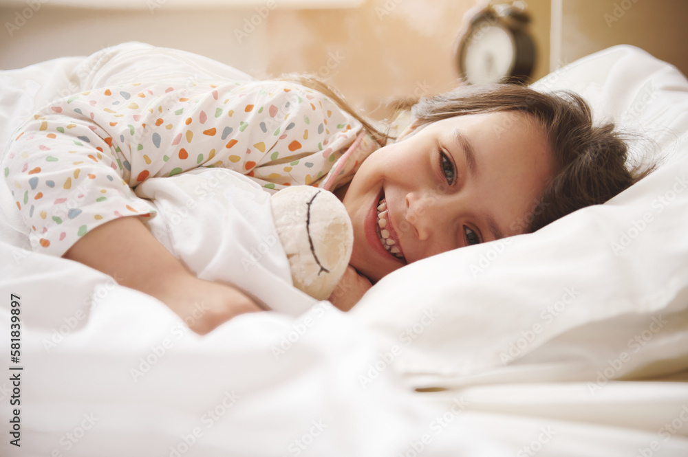 Adorable little child girl in pajamas, gently hugging her plush toy sheep, smiling looking at camera while awakening in bed at home in the morning, in the light children bedroom interior Adorable little child girl in pajamas, gently hugging her plush toy sheep, smiling looking at camera while awakening in bed at home in the morning, in the light children bedroom interior
