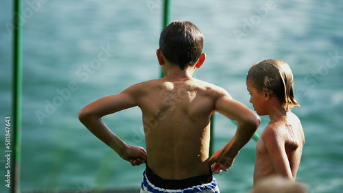 Back of two wet kids standing by lake dock. Young boy adjusting swim suit short. Children enjoying summer holiday vacations