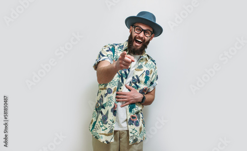 Ha ha, look at you! Portrait of a hipster guy in a Hawaiian shirt and hat laughing out loud and pointing his finger at the camera, making fun of a ridiculous appearance or a joke. 
