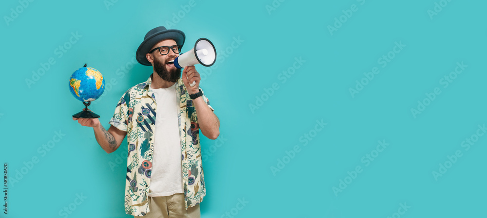 A young guy with a beard in a hat, Hawaiian shirt holds a globe and shouting to the side into a megaphone, isolated on a blue studio background. Travel agency concept - obrazy, fototapety, plakaty 