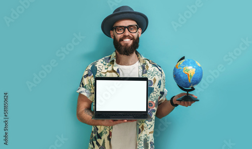 A young guy in a Hawaiian shirt with a beard holds a globe and a laptop in his hand demonstrating a blank screen with copy space for advertising and advertising text. travel agency concept