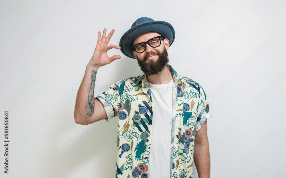 Fototapeta premium Some more, please. A cute hipster guy in a hat, with a beard gesticulates with his fingers, shows something small, asks for a little time, measures too small an object.