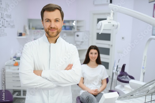 Male dentist in a room with medical equipment and patient on background.