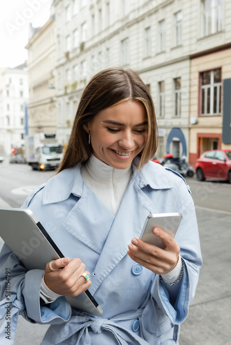 happy woman in blue trench coat using smartphone while holding laptop on street in Vienna.