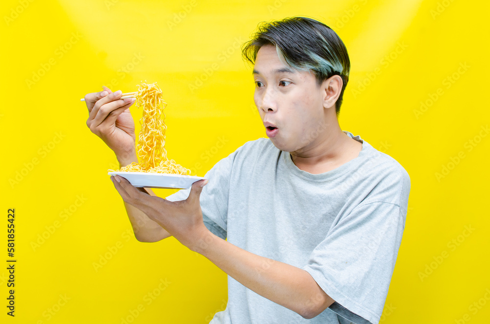 Happy euphoric asian young man in grey tshirt eating instant fried noodle using bamboo chopstick isolated over yellow background. stylish asian man with peek a boo hair eats instant noodle Happy euphoric asian young man in grey tshirt eating instant fried noodle using bamboo chopstick isolated over yellow background. stylish asian man with peek a boo hair eats instant noodle